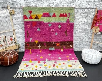 Puede incluir: Una alfombra marroquí colorida con patrones geométricos en tonos de rosa, verde, marrón y amarillo. La alfombra tiene un borde de flecos y está tejida con lana.