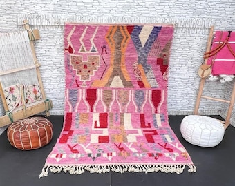 May include: A pink and white Moroccan rug with geometric patterns and fringe. The rug is displayed on a black floor with a brown leather pouf and a white pouf.