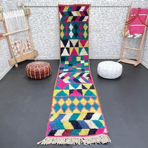May include: A colorful Moroccan runner rug with geometric patterns in shades of pink, blue, yellow, and green. The rug has a fringe on both ends.