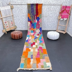 May include: A colorful Moroccan runner rug with a geometric pattern in shades of orange, pink, blue, green, and gray. The rug has a fringe border and is made of wool.