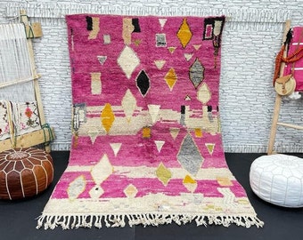 May include: A pink and white Moroccan rug with geometric patterns, including diamonds, triangles, and stripes. The rug has a fringe border and is displayed on a white tiled floor.