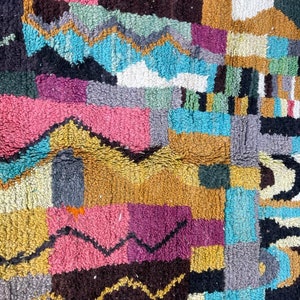 May include: A colourful Moroccan rug with a geometric pattern in shades of blue, pink, yellow, brown, and grey. The rug is made of wool and has a soft, fuzzy texture.