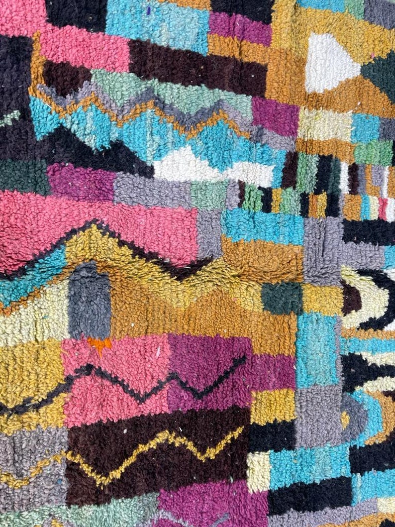 May include: A colourful Moroccan rug with a geometric pattern in shades of blue, pink, yellow, brown, and grey. The rug is made of wool and has a soft, fuzzy texture.