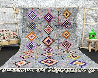 May include: A gray Moroccan rug with a geometric pattern in shades of pink, blue, orange, and green. The rug has a fringe border and is woven with a traditional Moroccan design.
