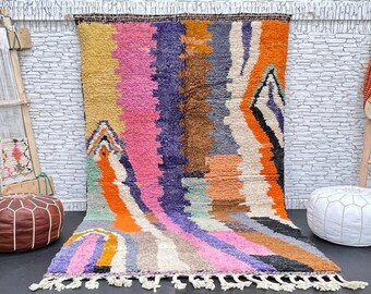 May include: A colorful Moroccan rug with a geometric pattern in shades of pink, orange, yellow, blue, and brown. The rug has a fringe border and is displayed on a white brick wall with a wooden loom and two poufs.