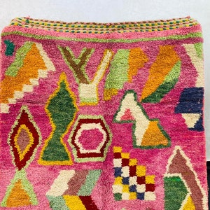 May include: A pink rug with a geometric pattern in shades of orange, yellow, green, blue, and brown. The rug has a fringe border.