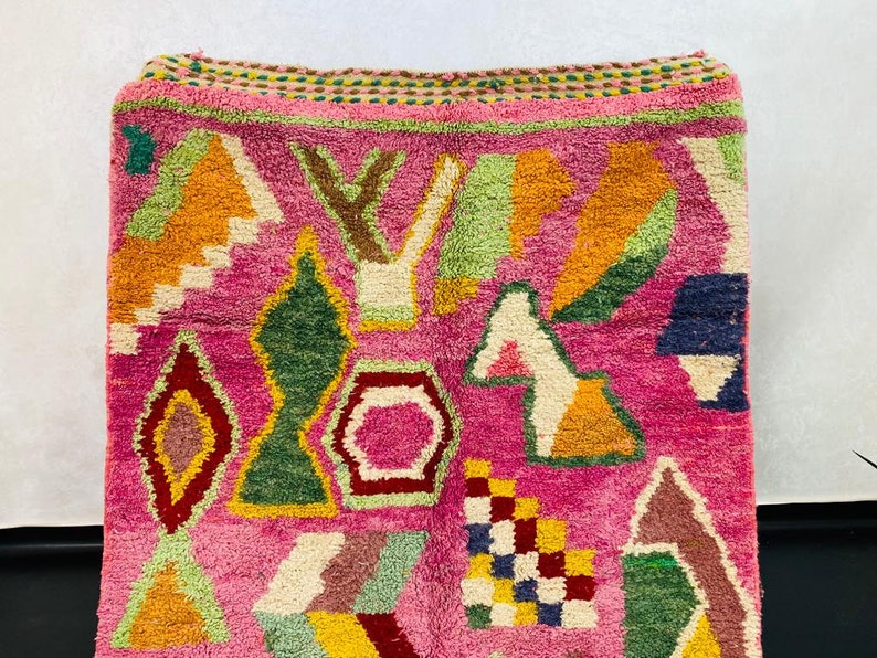 May include: A pink rug with a geometric pattern in shades of orange, yellow, green, blue, and brown. The rug has a fringe border.