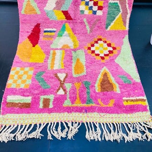 May include: A pink rug with a geometric pattern in shades of green, yellow, orange, brown, and white. The rug has a fringe border.