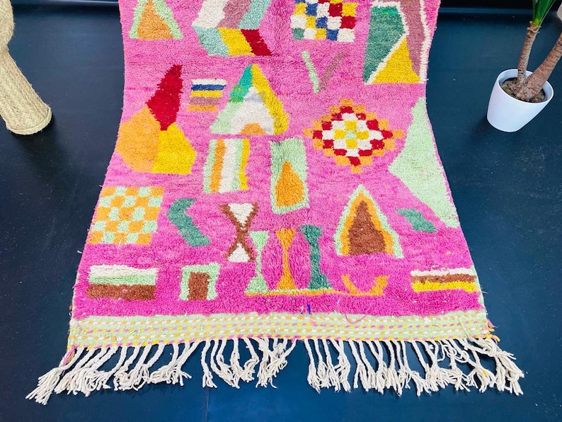 May include: A pink rug with a geometric pattern in shades of green, yellow, orange, brown, and white. The rug has a fringe border.