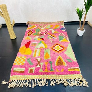 May include: A pink and multi-colored Moroccan rug with geometric patterns and fringe. The rug is made of wool and has a hand-woven texture.