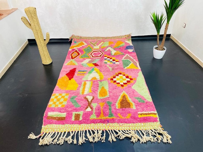May include: A pink and multi-colored Moroccan rug with geometric patterns and fringe. The rug is made of wool and has a hand-woven texture.