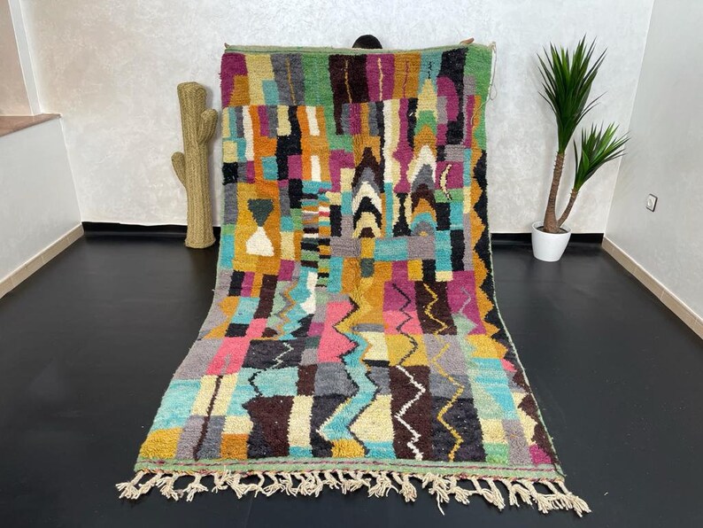 May include: A colourful Moroccan rug with geometric patterns in shades of pink, blue, green, yellow, and brown. The rug has a fringe border and is made of wool.
