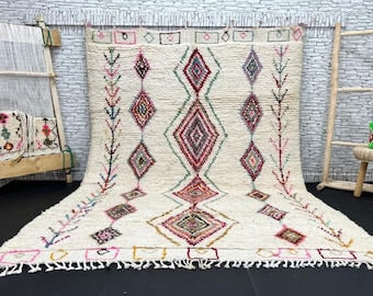 May include: A white Moroccan rug with a diamond pattern in shades of pink, red, green, and blue. The rug has a fringe border and is woven with a variety of textures.