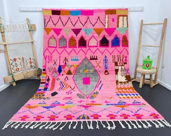May include: A pink Moroccan rug with a geometric pattern in shades of pink, blue, orange, and yellow. The rug has a fringe border and is woven with a variety of textures.