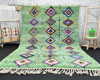 May include: A green Moroccan rug with a geometric pattern in shades of pink, blue, and yellow. The rug has a fringe border and is made of wool.