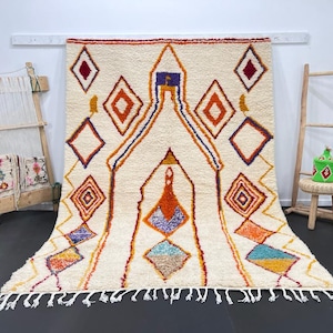 May include: A white Moroccan rug with a geometric pattern in shades of orange, red, purple, and blue. The rug has a fringe border and is hanging on a wall.