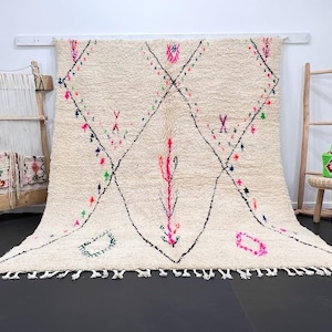 May include: A white Moroccan rug with a diamond pattern and colorful accents. The rug has a fringe border and is hanging on a wall.