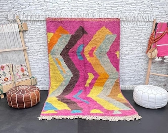 May include: A colorful Moroccan rug with a geometric pattern in shades of pink, orange, yellow, blue, and brown. The rug has a fringe border and is laid out on a gray floor.