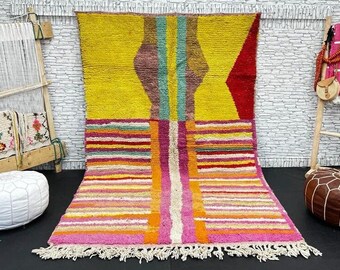 May include: A colorful Moroccan rug with a geometric pattern in shades of yellow, pink, red, and orange. The rug has a fringe border and is woven with a variety of textures.