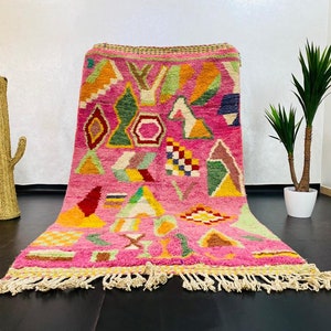 May include: A pink Moroccan rug with a geometric pattern in shades of green, yellow, red, and blue. The rug has a fringe border and is made of wool.