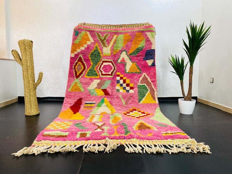 May include: A pink Moroccan rug with a geometric pattern in shades of green, yellow, red, and blue. The rug has a fringe border and is made of wool.