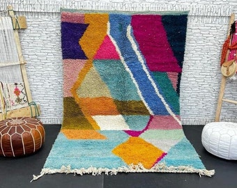 May include: A colorful abstract Moroccan rug with geometric shapes in shades of blue, pink, orange, and green. The rug has a fringe border and is displayed on a black floor with two poufs.