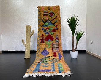 May include: A long, rectangular rug with a geometric pattern in shades of yellow, red, blue, green, and purple. The rug has a fringe border and is made of wool.