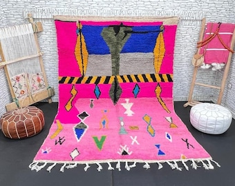 Puede incluir: Una alfombra marroquí rosa con un patrón geométrico en tonos de azul, amarillo, verde y negro. La alfombra tiene un borde de flecos y está hecha de lana.