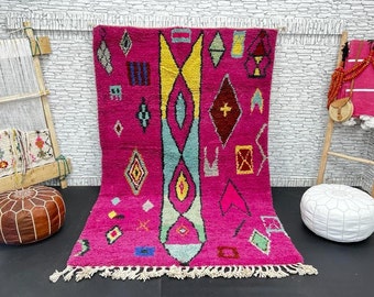 Puede incluir: Una alfombra marroquí rosa con un patrón geométrico en tonos de amarillo, azul, rojo y verde. La alfombra tiene un borde de flecos y se muestra sobre un suelo negro.