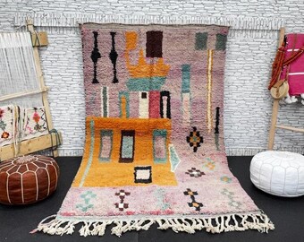 Puede incluir: Una alfombra marroquí colorida con patrones geométricos en tonos de rosa, naranja, azul y verde. La alfombra tiene un borde de flecos y se muestra en una pared de ladrillo blanco con un telar de madera y dos pufs.