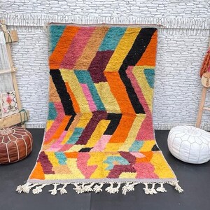 May include: A colourful Moroccan rug with a geometric pattern in shades of orange, yellow, pink, blue, and black. The rug has a fringed border and is laid out on a dark floor.