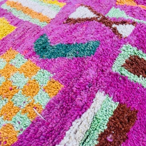May include: A close-up of a colorful Moroccan rug with a pink background and geometric patterns in shades of green, orange, yellow, and white.