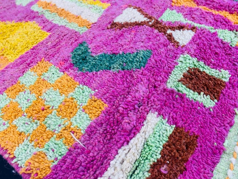 May include: A close-up of a colorful Moroccan rug with a pink background and geometric patterns in shades of green, orange, yellow, and white.