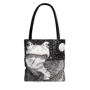 May include: Black and white tote bag featuring a detailed illustration of a frog in a nighttime scene. The design includes a starry sky, a full moon, and water. The bag has black handles.