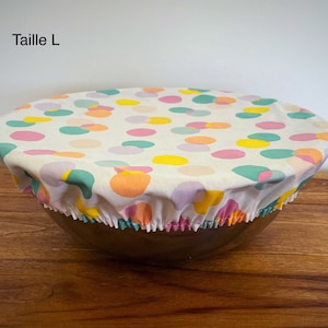 Op de afbeelding: Een witte stoffen komdeksel met een kleurrijk stippenpatroon in roze, geel, oranje en groen. De deksel ligt op een houten oppervlak en de tekst "Taille L" is zichtbaar in de linkerbovenhoek.