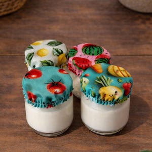 May include: Four small glass jars filled with a white substance, each with a patterned fabric lid. The lids display designs of lemons, watermelons, tomatoes, and vegetables. The jars are on a wooden surface, with plants in the background.