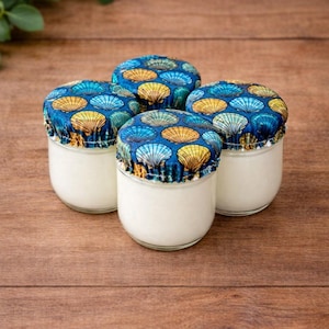 Puede incluir: Cuatro pequeños frascos de vidrio transparente con contenido blanco, cubiertos con tapas de tela. Las tapas presentan un fondo azul con un patrón de conchas marinas amarillas y azules. Los frascos están sobre una superficie de madera.