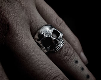 May include: A silver skull ring worn on a finger. The ring is detailed with a textured skull design.