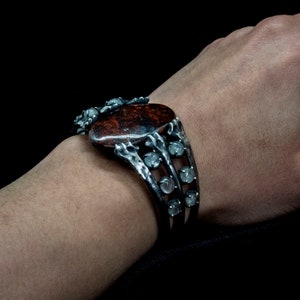 May include: A silver bracelet with a large, oval-shaped red gemstone surrounded by smaller, clear gemstones. The bracelet has a unique, organic design with a textured finish.