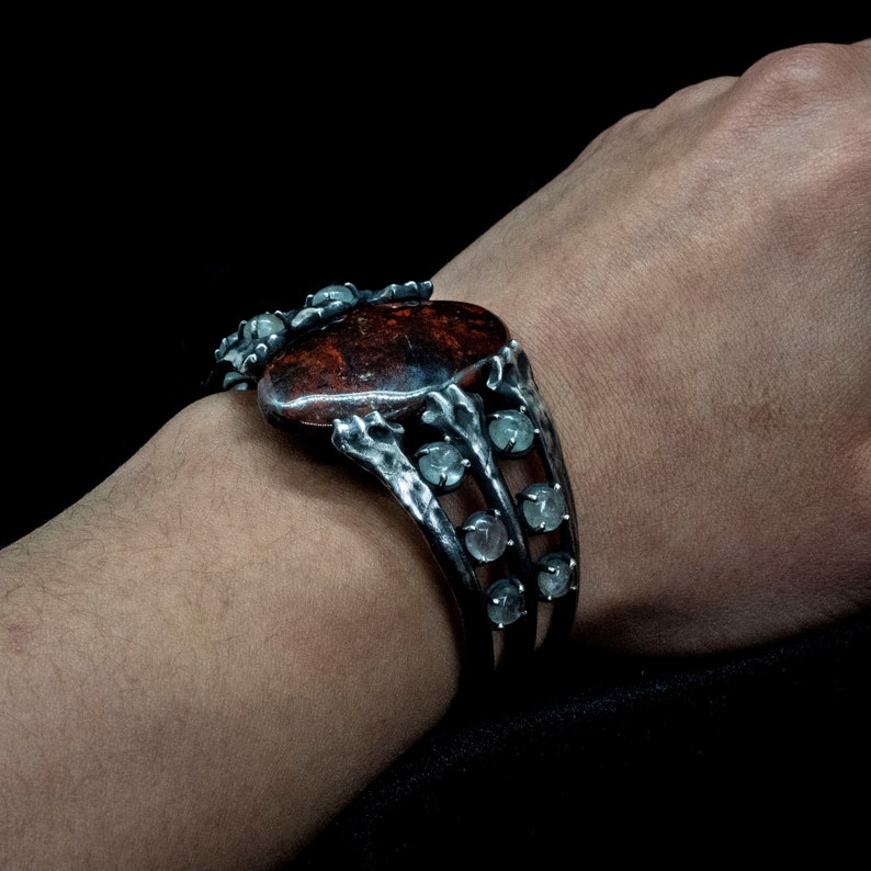 May include: A silver bracelet with a large, oval-shaped red gemstone surrounded by smaller, clear gemstones. The bracelet has a unique, organic design with a textured finish.