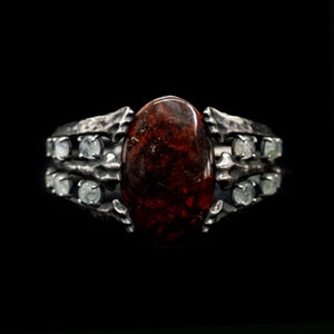 May include: A silver ring with a large, oval-shaped red gemstone surrounded by smaller, clear gemstones.