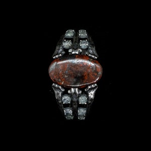 May include: A silver ring with a red and brown stone set in a skeletal design. The ring is adorned with clear gemstones.