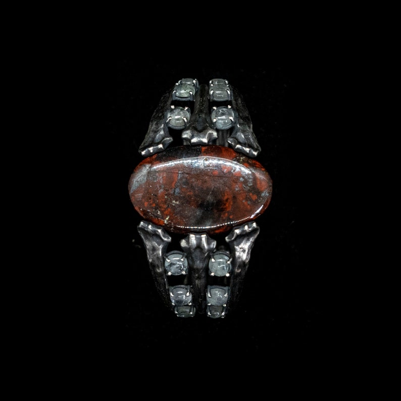 May include: A silver ring with a red and brown stone set in a skeletal design. The ring is adorned with clear gemstones.