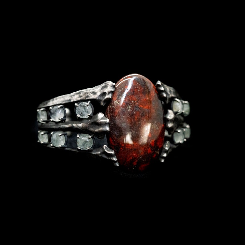 May include: A silver ring with a large, oval-shaped red gemstone surrounded by smaller, clear gemstones.