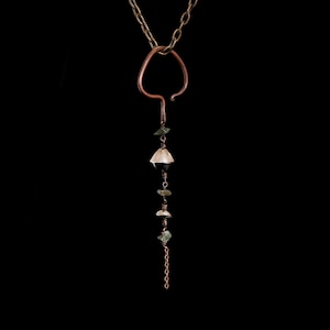 May include: A handcrafted copper pendant necklace with a unique, abstract design. The necklace features a copper chain and a pendant with a teardrop shape, adorned with green and clear gemstones and a black bead. The necklace hangs against a black background.