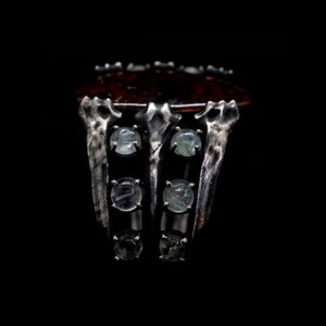 May include: A silver ring with a large, dark red stone set in the center. The ring has a unique, claw-like design with six smaller, clear stones set around the main stone.