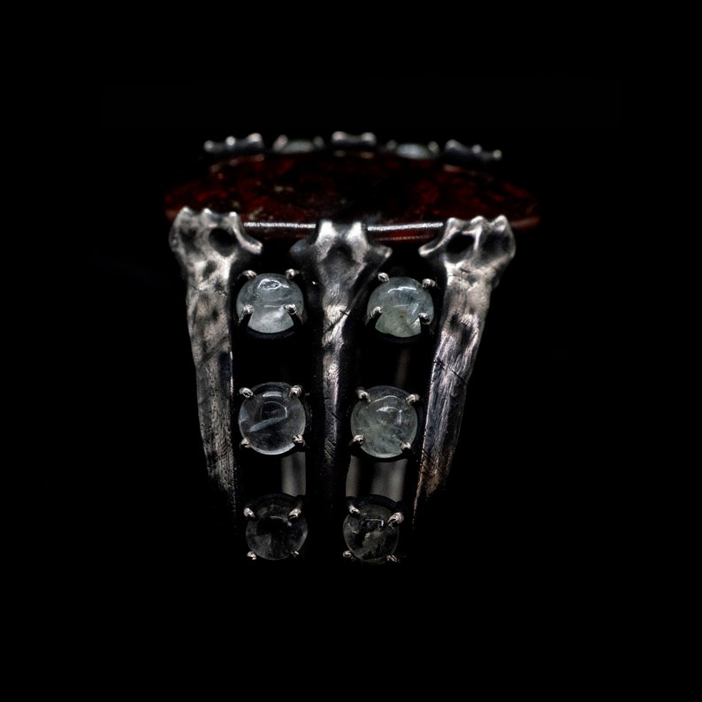 May include: A silver ring with a large, dark red stone set in the center. The ring has a unique, claw-like design with six smaller, clear stones set around the main stone.