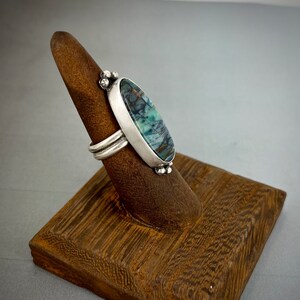May include: A silver ring featuring an elongated oval stone with dark blue, green, and brown veining. The stone is set in a smooth silver bezel with small decorative silver spheres at the top and bottom. The band consists of two parallel silver wires.