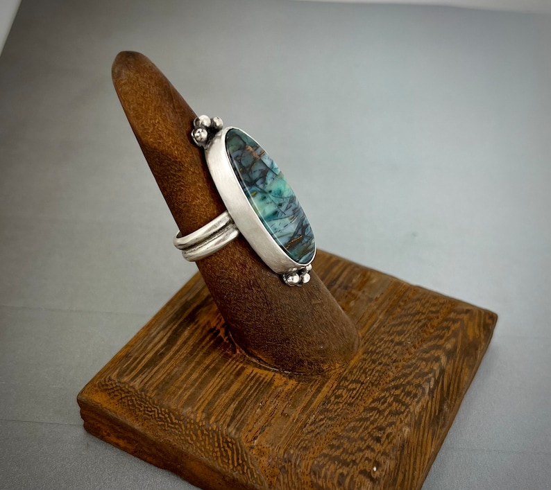 May include: A silver ring featuring an elongated oval stone with dark blue, green, and brown veining. The stone is set in a smooth silver bezel with small decorative silver spheres at the top and bottom. The band consists of two parallel silver wires.