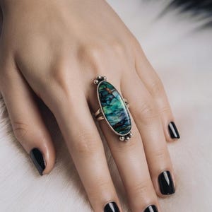 May include: A sterling silver ring featuring an elongated oval-shaped stone with swirling patterns of deep green, teal, and dark brown. The metal setting includes small decorative silver spheres at the top and bottom of the stone, with a split band.